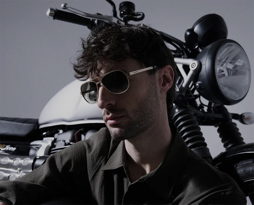 Modello che indossa gli occhiali Serpico sole di Emblema ed è seduto davanti ad una moto triumph.